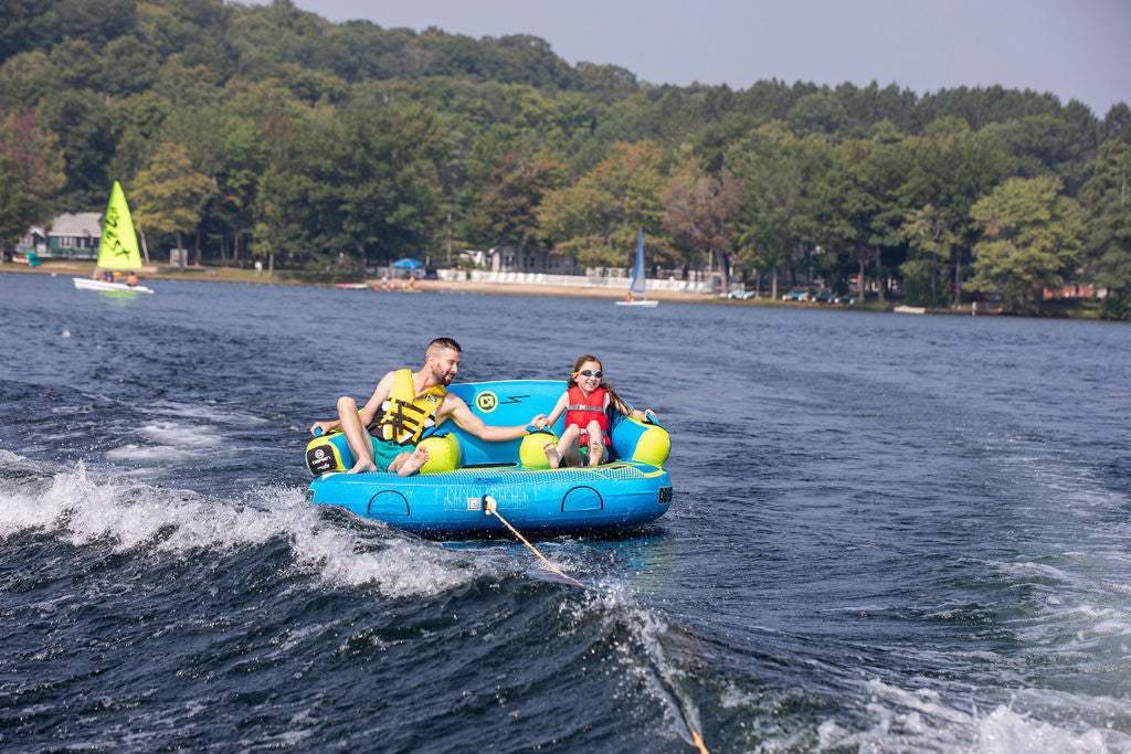 An adult and a child, both wearing life jackets, are riding on a water tube in a lake. The adult is holding the child's hand.