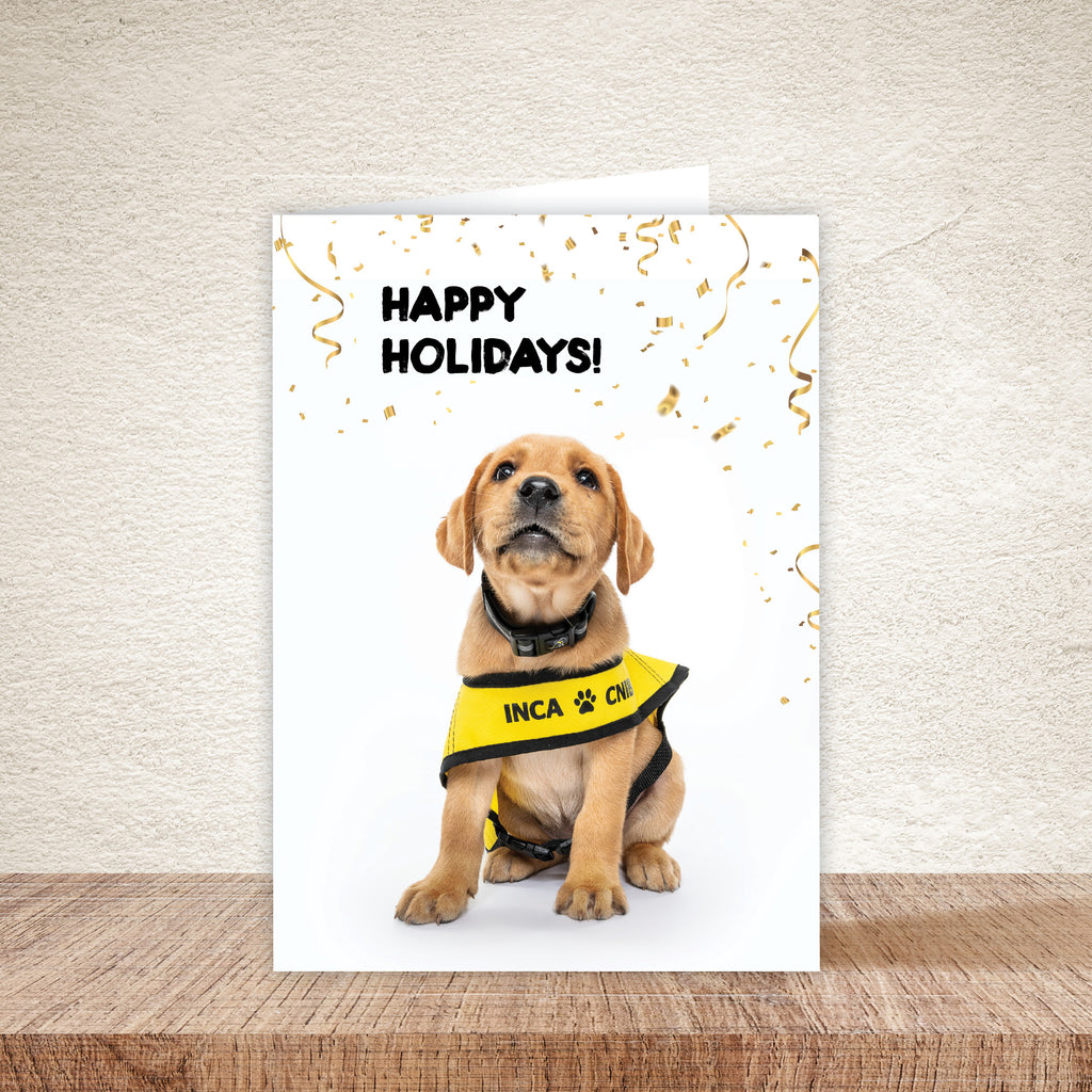 Holiday card featuring a yellow Labrador puppy in a yellow vest with the text ‘Happy Holidays!’ at the top, displayed on a wooden surface against a beige wall background.