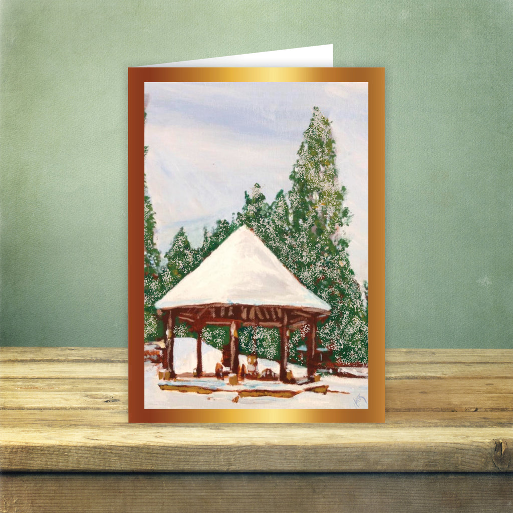 Holiday card with an illustrated winter scene of a pavilion and snow-covered trees, displayed on a wooden surface against a green wall background.