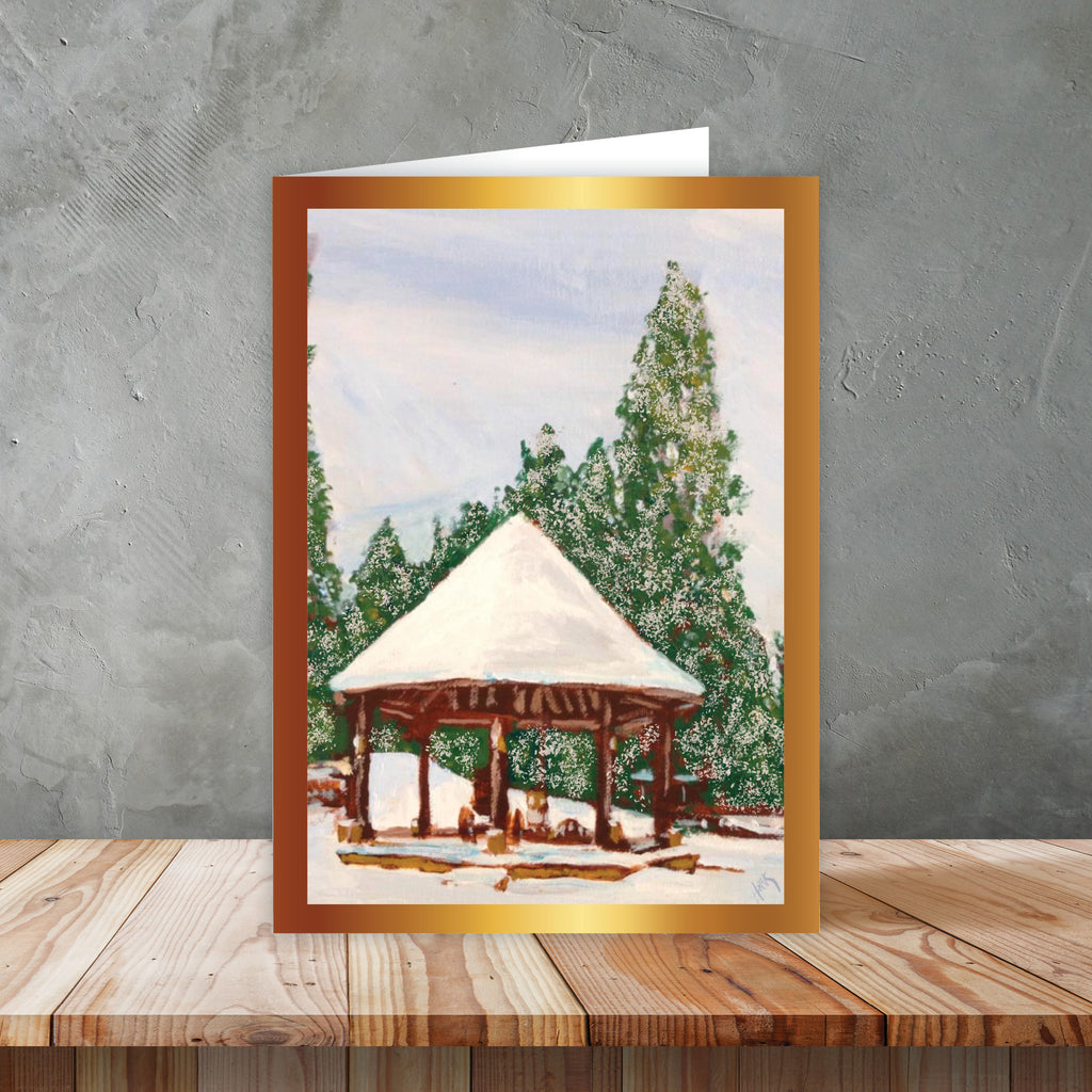 Holiday card with an illustrated winter scene of a pavilion and snow-covered trees, displayed on a wooden surface against a gray wall background.
