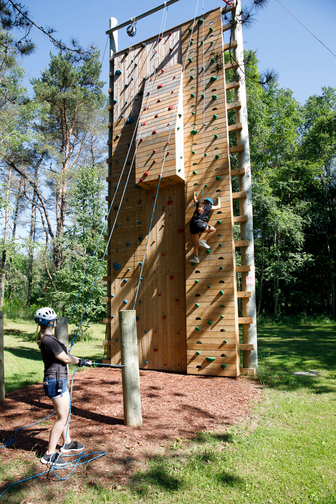 A participant, wearing a helmet and harness, climbs an outdoor wall with colourful holds and ropes on a sunny summer day. On the ground, another helmeted participant holds the climbing rope. Greenery fills the background.
