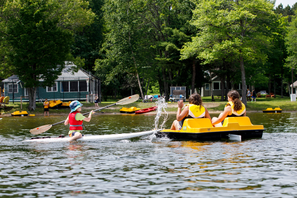 Two people in life jackets paddle a boat, their backs to the camera, while a kid on a nearby paddleboard splashes them with a paddle. The adults try to shield themselves from the water. In the background, there’s a small beach with parked paddle boats and kayaks, and houses visible behind the trees.