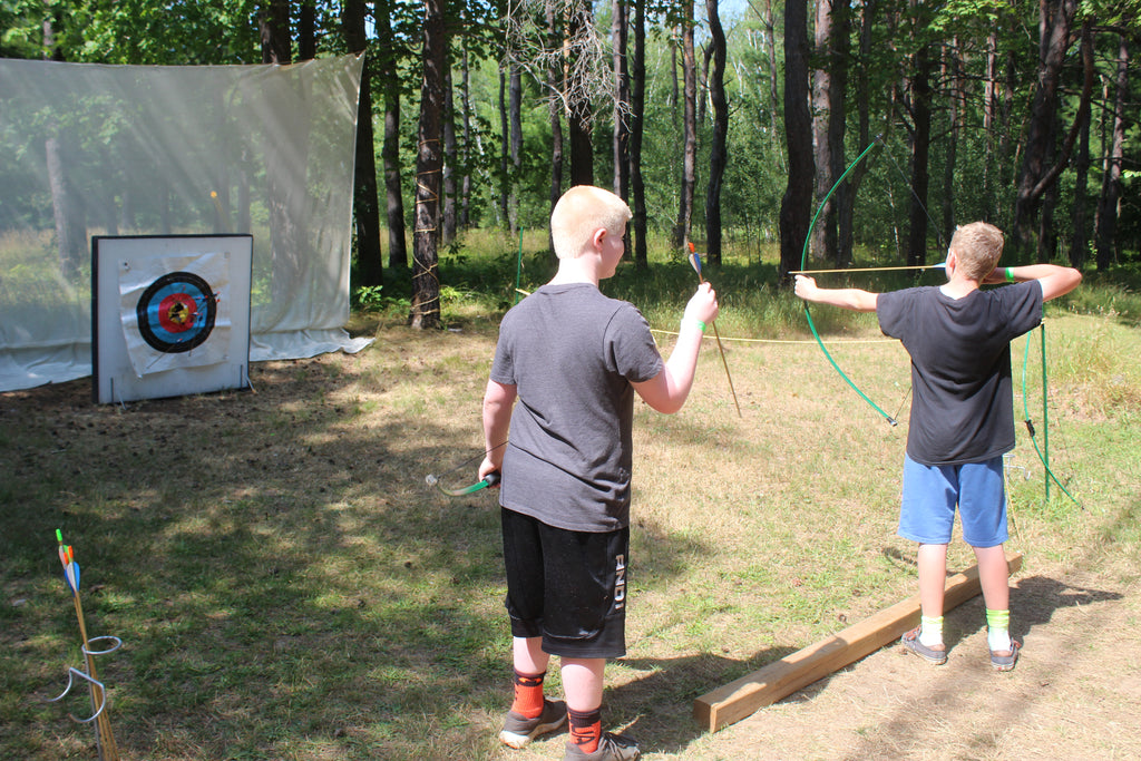 A young boy with light skin aims his bow while practicing archery outdoors, as another boy with light skin watches nearby. Tall green trees fill the background.
