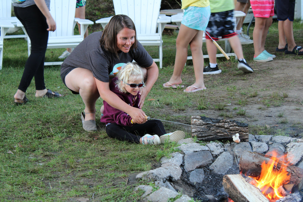 A woman with light skin and her young daughter roast a marshmallow on a stick over a campfire, with other campers gathered in the background.